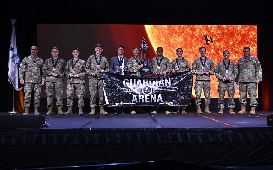 11 men, all but one in uniform, pose for a photo on a stage while those in the center of the line hold a poster reading “Guardian Arena.”