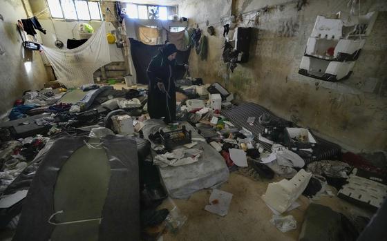 A woman in a black hijab walks through a prison cell strewn with dirty mattresses, clothes and blankets.