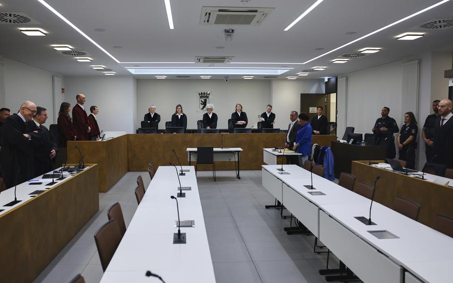 A group of people in professional attire stand behind desks that line the perimeter of a courtroom.