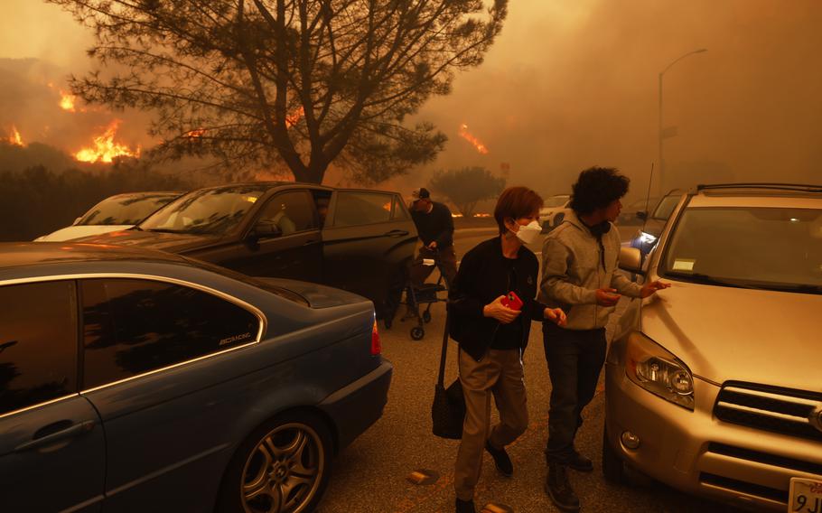 People rush to cars on a street as a wildfire burns in hills in the background.