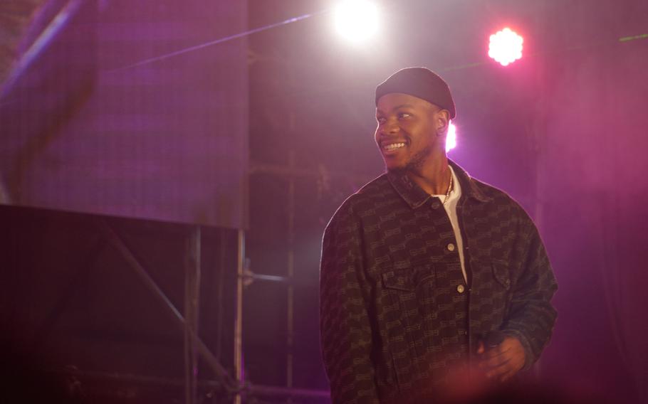 A man wearing a beanie and dark jacket smiles while he is backlit with pink stage lights.