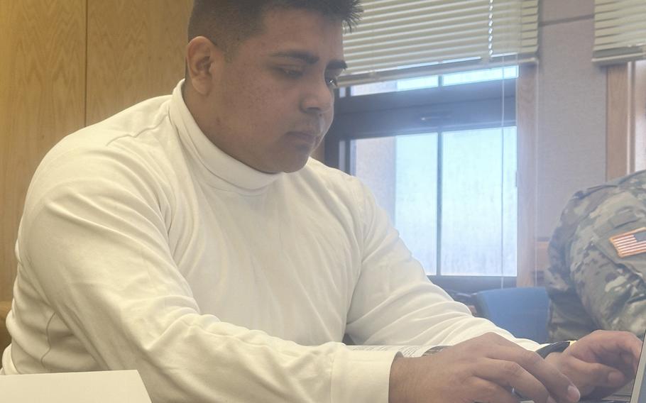 A man in a white turtleneck shirt sits at a table and types on a laptop computer.