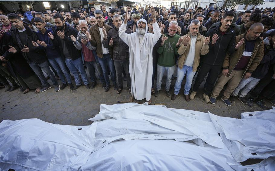 A man in religious attire leads a line of mourners in prayer as they stand in front of bodies covered by sheets.