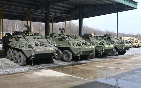 A row of four green armored vehicles rest under a roof, with other military vehicles in the background.