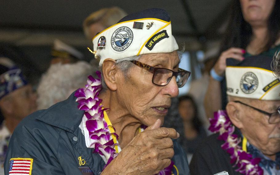 An elderly man wearing a hat and a flowered lei sits and speaks.