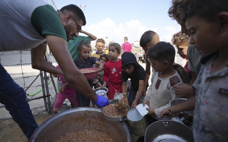 A man spoons food onto the dishes of displaced Palestinian children gathered around a large pot.