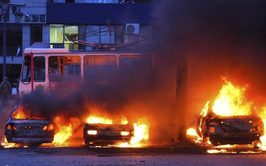 Three cars are seen burning and emitting smoke in front of a bus.