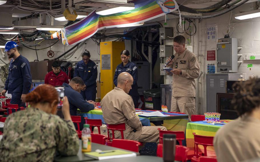 A handful of service members seated at a table while a sailor stands and speaks into a microphone below a rainbow flag.