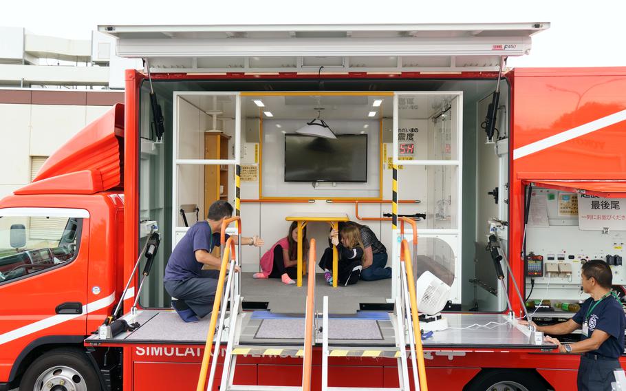 A mobile earthquake simulator on an orange truck contains a room.  Two kids and a mom huddle under the table in the room as two workers look on. 