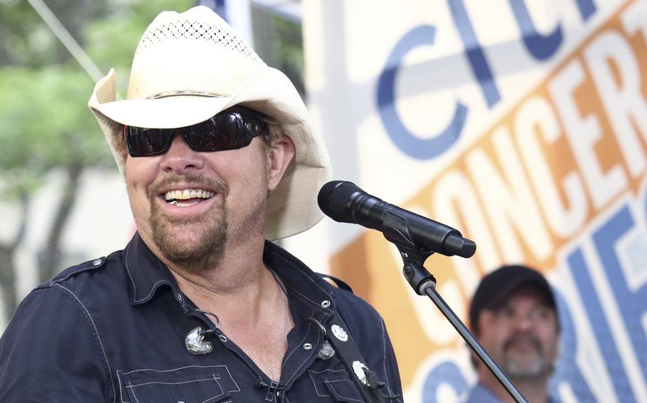 Toby Keith, wearing a white cowboy hat and dark sunglasses, flashes a smile.