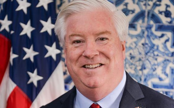 A man with white hair wearing a dark suit and red tie poses in front of a U.S. flag.