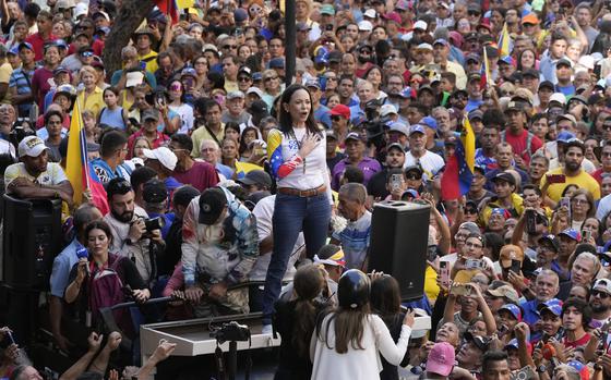 A woman in a white shirt and blue jeans stands on a pedastal with her hand over he heart while surrounded by supporters waving Venezuelan flags.