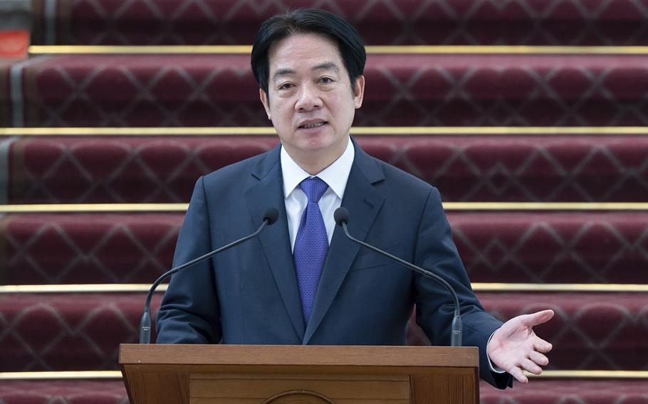 A Taiwanese man in a dark suit stands at a wooden podium and speaks into two microphones in front of a red-carpeted staircase.