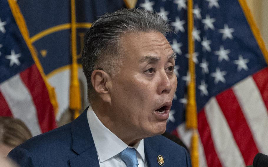 A congressman wearing a blue suit speaks into a microphone while seated in front of U.S. flags during a hearing.