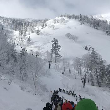 A line of skiers and snowboarders trails down a hill in the foreground with a view of a snowy mountain in the background.