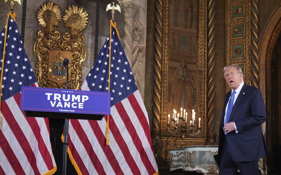 An older man in a suit and blue tie walks towards a podium flanked by American flags and set in front of a wall filled with ornate gold frames and a candelabra.