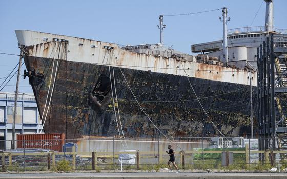 A person runs on a path alongside a large, rusty ship with United States written on it.