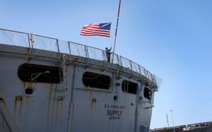 A mariner aboard the fast combat support ship USNS Supply raises the national ensign as the ship moors in Naval Station Norfolk, Va., July 27, 2024, following an eight-month deployment in support of the USS Dwight D. Eisenhower Carrier Strike Group. The Navy is looking for ways to address a critical shortage of qualified civilian mariners without sidelining 17 of its logistics ships. 