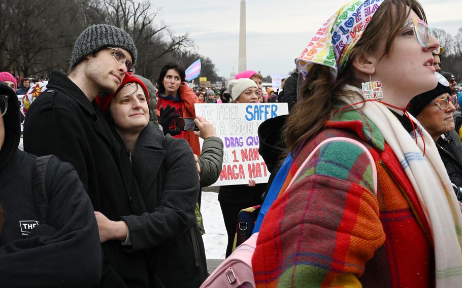 A protesting couple embraces on the left while another protestor on the right looks forward; the Washington Monument is seen in the distance in the center of the photo.