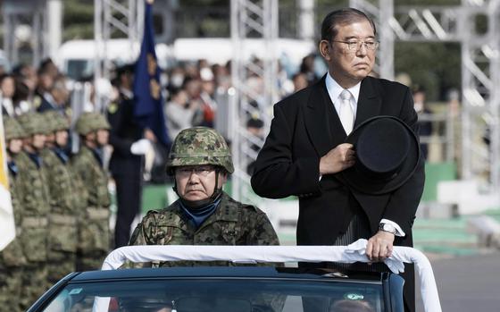 Japanese Prime Minister Shigeru Ishiba stands in the back seat of a car next to a seated soldier during military parade.