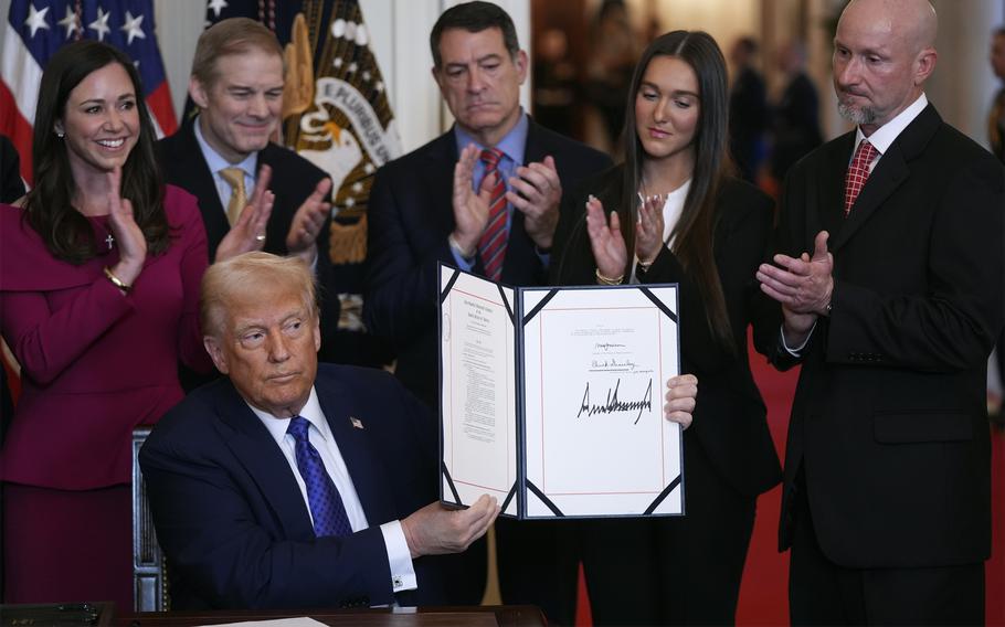 Donald Trump sits at a desk and holds up a folder with a signed bill while supporters stand behind him and clap.