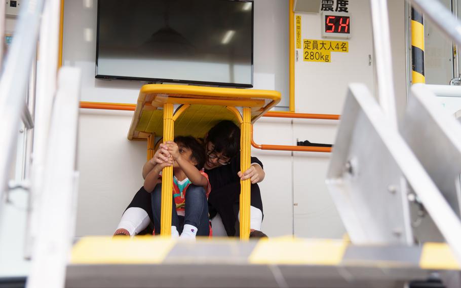 A woman and a boy huddle under a yellow table in an earthquake simulator. 