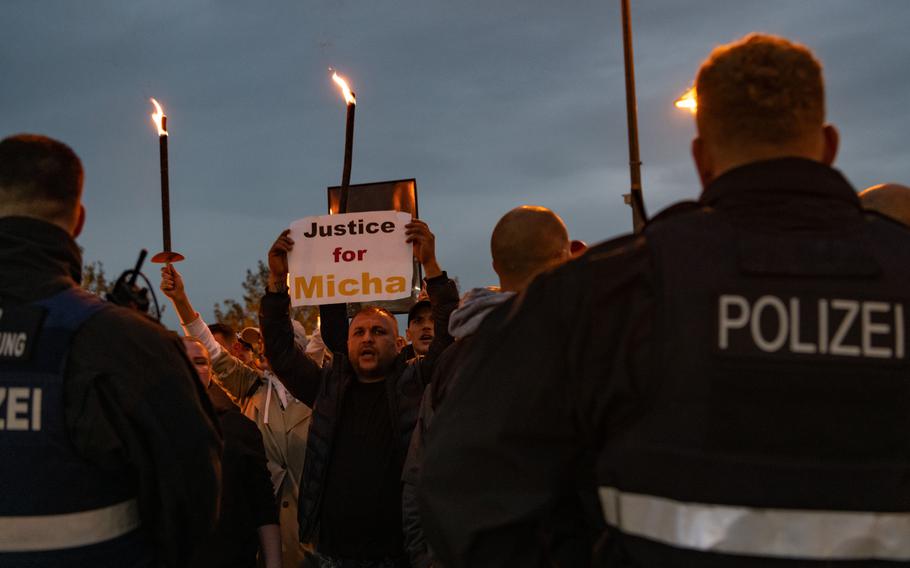 German police face protesters outside Spangdahlem Air Base in the evening. A man holds a sign that says “Justice for Micha.” Some protesters hold torches.