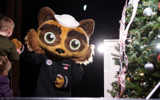 A mascot with a large head of a tanuki Japanese raccoon dog and wearing a Navy uniform greets children next to a Christmas tree.