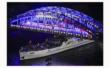 Athletes travel by boat down the Seine River in Paris, France, during the opening ceremony of the 2024 Summer Olympics, on July 26, 2024. On the bridge above the boat, DJ and LGBTQ+ icon Barbara Butch surrounded by drag artists, dancers and others, gets a party going.