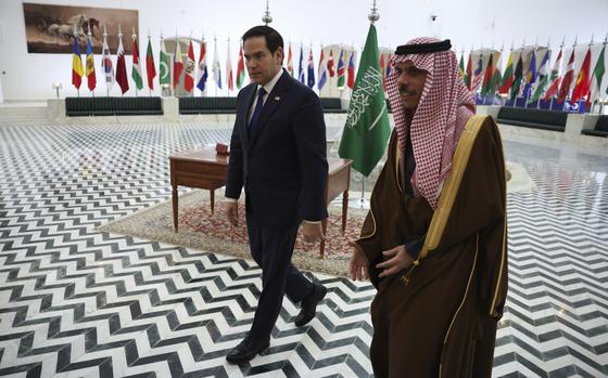 Marco Rubio and Prince Faisal Farhan Al Saud walk through a room with national flags in the background.