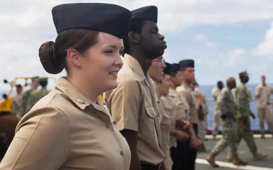 A female Navy sailor in dress uniform stands at attention next to other sailors on the deck of a ship.