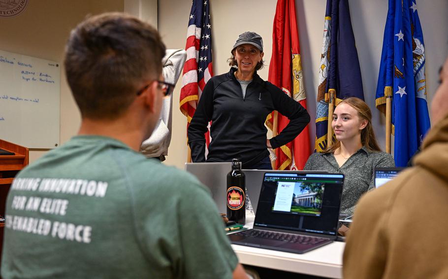 A service member in a baseball cap talks to cadets sitting in front of computers. 