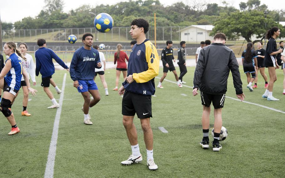 High school soccer players in warm-up jackets and shorts dribble soccer balls on a field.