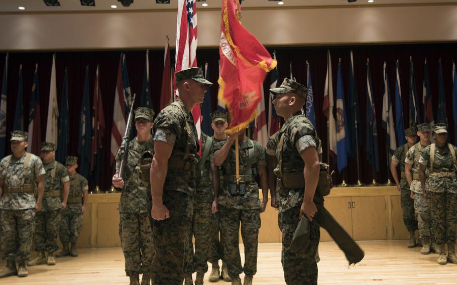 Two Marine Corps officers stand at attention facing each other in front of Marines holding flags.