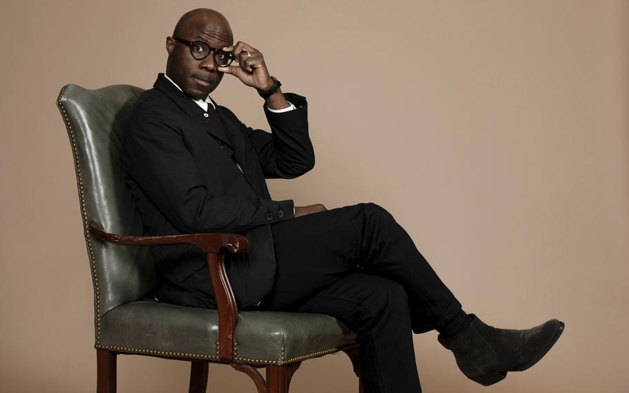 Filmmaker Barry Jenkins, in a black suit and black boots, sits in a chair and pinches the left lens of his eyeglasses with two fingers of his left hand.