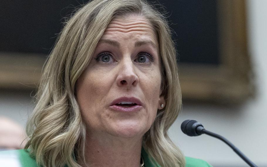 A close-up view of a woman with blonde hair and a green suit speaking into a microphone during a congressional hearing.