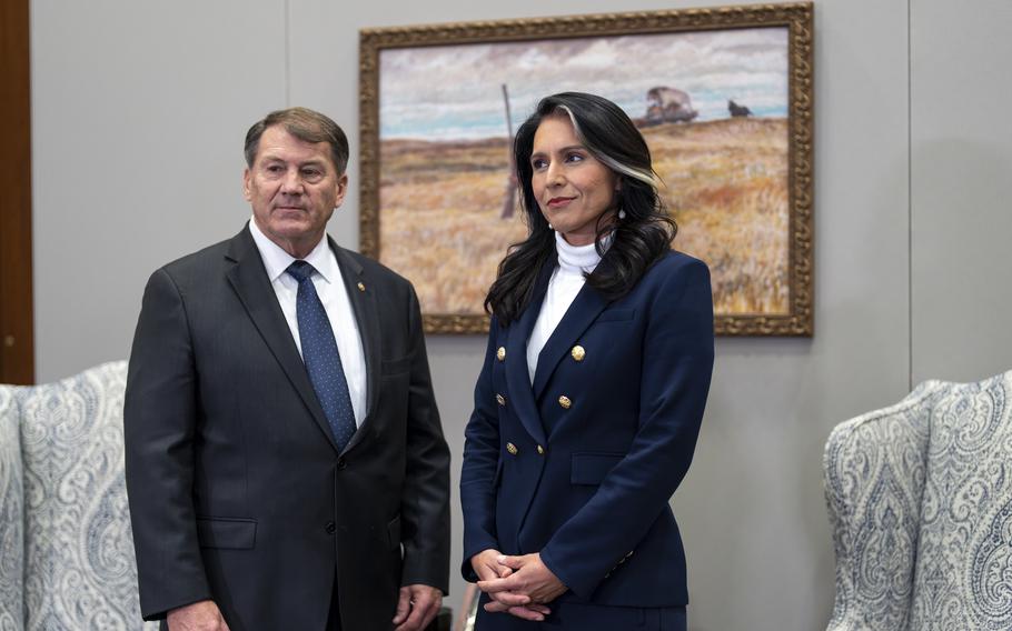 A woman in a turtleneck and navy suit stands with a man in a gray suit with a navy tie in front of a painting of a wheat field.