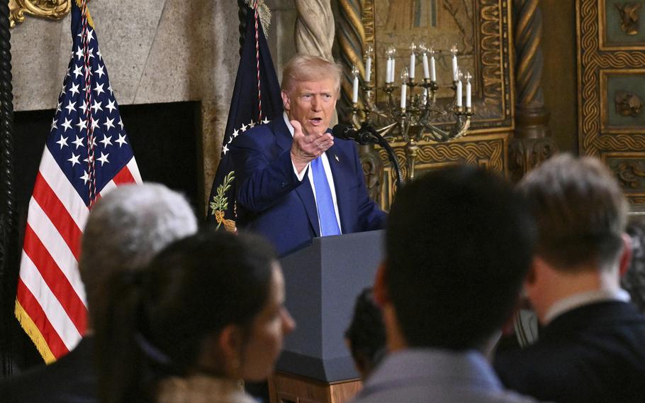 President Trump speaks at a podium while raising a hand.