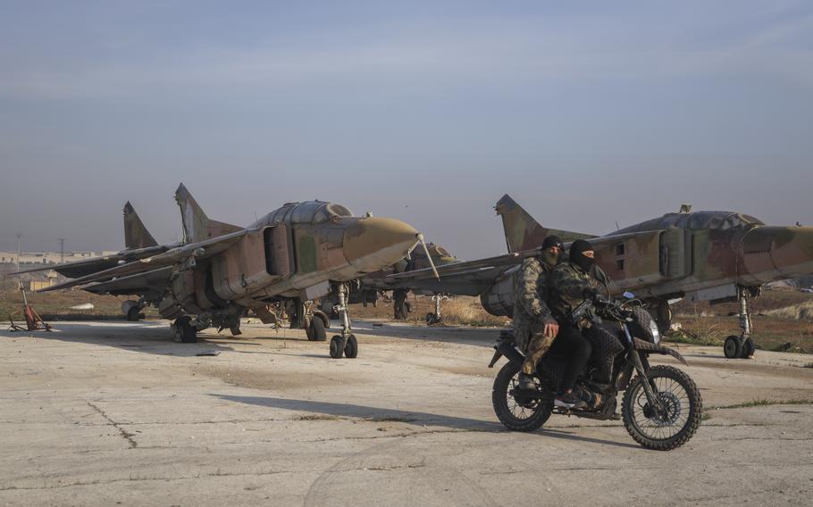 Two men in military gear and with their faces covered ride a motorcycle in front of Syrian army planes.