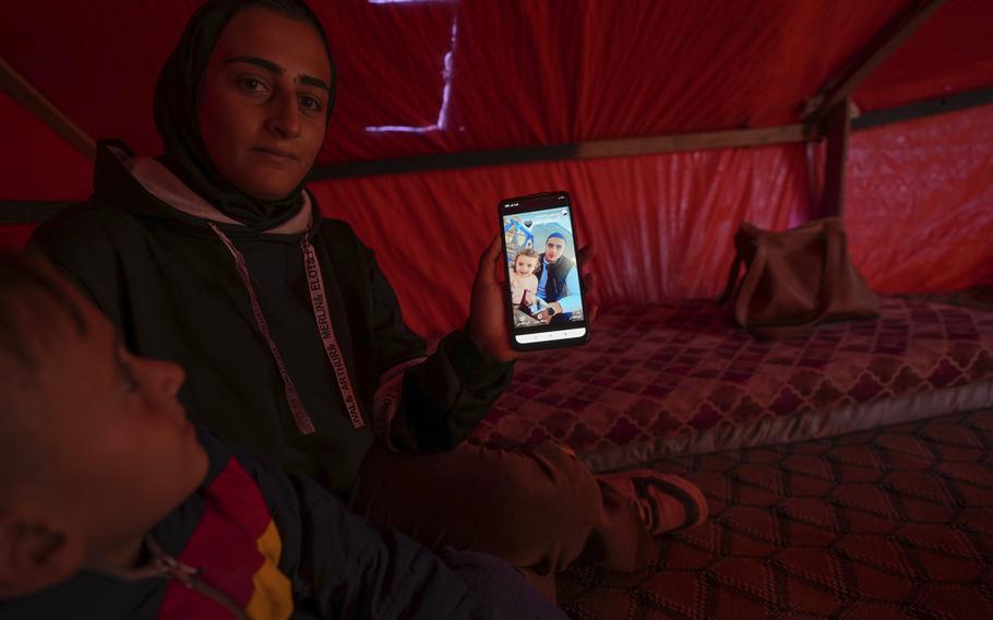 A woman wearing a head covering and her young son sit inside a tent and hold up a phone with a picture of a man and a young girl.