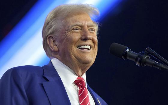 A man wears a blue suit and a red striped tie and smiles as he stands in front of a microphone.