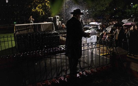 A rabbi, silhouetted by a spotlight, stands at a podium next to a raised coffin and delivers a eulogy to a crowd of mourners.