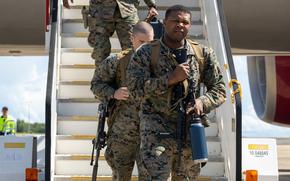 Marines in camouflage uniforms carrying machine guns descend the steps off an airplane onto a runway.