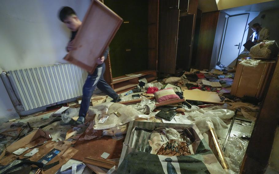 A man carries a rectangular board through a room littered in broken art frames, pillows and trash.