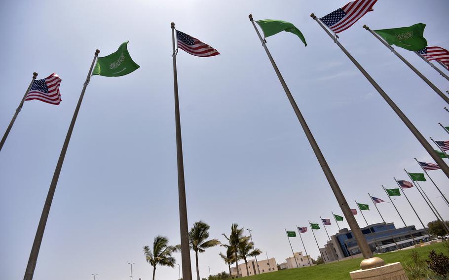 Saudi and U.S. flags, seen from below, stand in a ring around a grassy plaza.