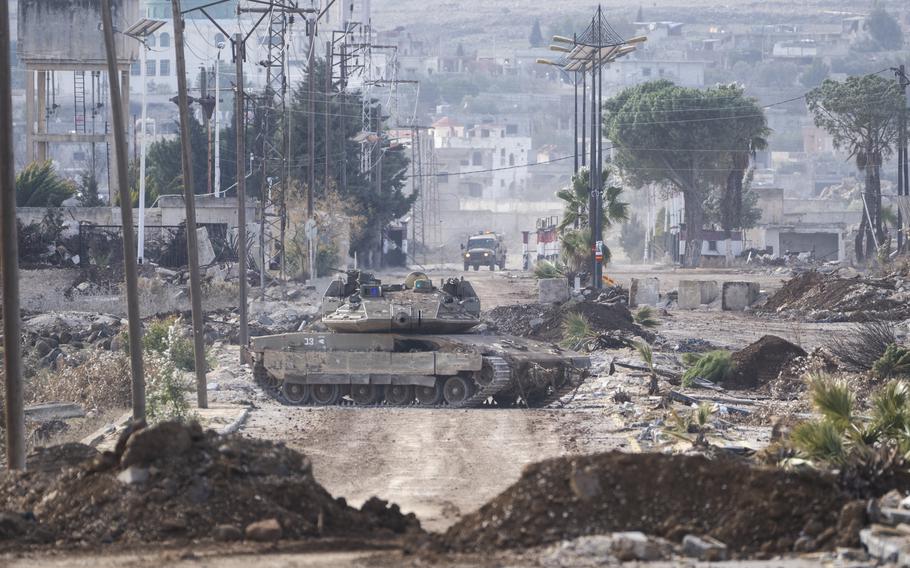 A road with debris on either side leads into a town in the distance, with a tank blocking the road in the center of the photo.