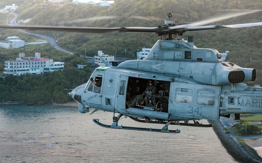 A helicopter with an open side door flies over water with coastal buildings in the background.