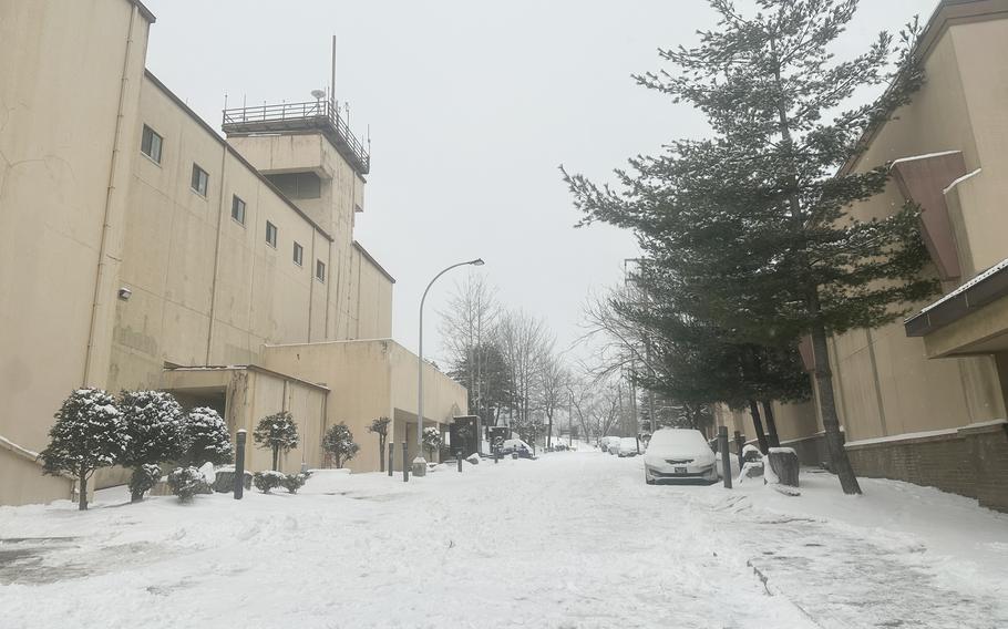 Snow covers a street and parked cars next to military buildings.