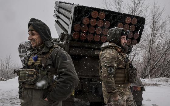 Two soldiers in combat gear stand next to an armored vehicle with a mounted rocket launcher.