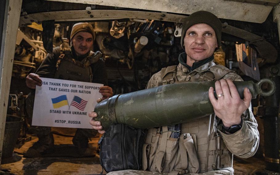 A Ukraine soldier holds a sign that says “USA thank you for the support that saves our nation,” “Stand with Ukraine” and “#Stop_Russia.” A second soldier holds a mortar shell.